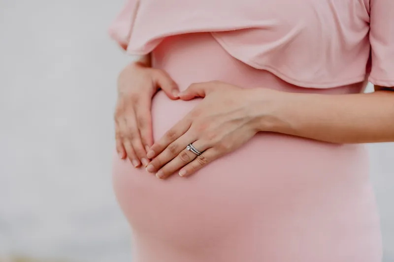 Zwangere vrouw in een roze jurk vormt een hartje met haar handen op haar buik